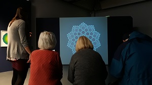 A group of three middle-aged women and their art tutor, a young woman, sketch in front of a geometric light projection in the gallery